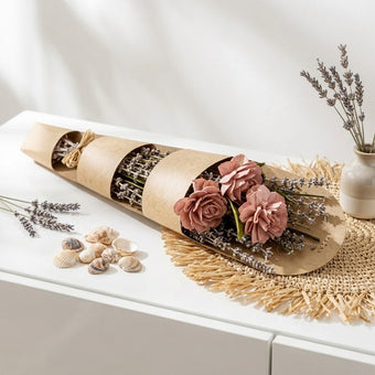 Buchet decorativ cu flori uscate roz și crenguțe de lavandă în ambalaj din hârtie kraft, așezat pe o comodă albă lângă scoici decorative.