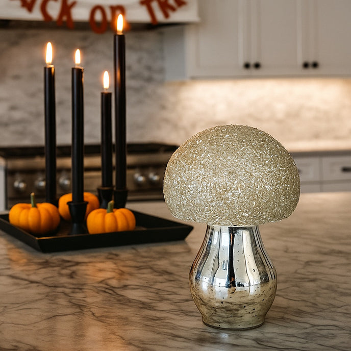 Ciupercă decorativă din sticlă, de culoare argintie și pălărie cu paiete albe, așezată pe un blat gri de bucătărie, decorată de Halloween
