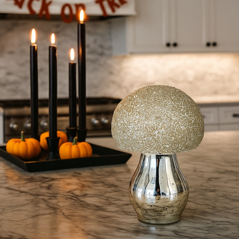 Ciupercă decorativă din sticlă, de culoare argintie și pălărie cu paiete albe, așezată pe un blat gri de bucătărie, decorată de Halloween