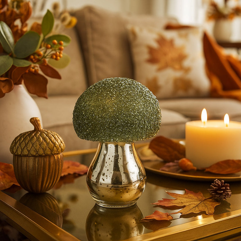 Ciupercă decorativă din sticlă, de culoare argintie și pălărie cu paiete verzi, așezată pe o masă decorată de toamnă