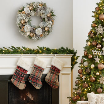 Coroniță de Crăciun cu conuri ninse și globuri aurii, agățată pe un perete alb, deasupra unui șemineu alb decorat de Caăciun