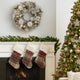 Coroniță de Crăciun cu conuri ninse și globuri aurii, agățată pe un perete alb, deasupra unui șemineu alb decorat de Caăciun