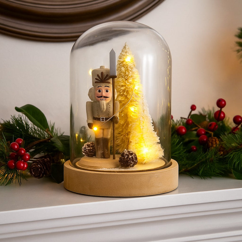 Decorațiune spărgător de nuci în clopot de sticlă cu lumini led așezată pe o manta de șemineu, albă, decorată de Crăciun