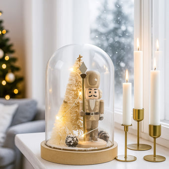 Decorațiune spărgător de nuci în clopot de sticlă cu lumini led așezată pe un pervaz alb, lângă 3 lumânări aprinse