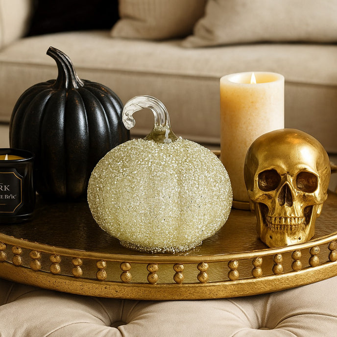 Dovleac decorativ din sticlă, de culoare albă și acoperit cu paiete, așezat pe o tavă aurie decorată de Halloween