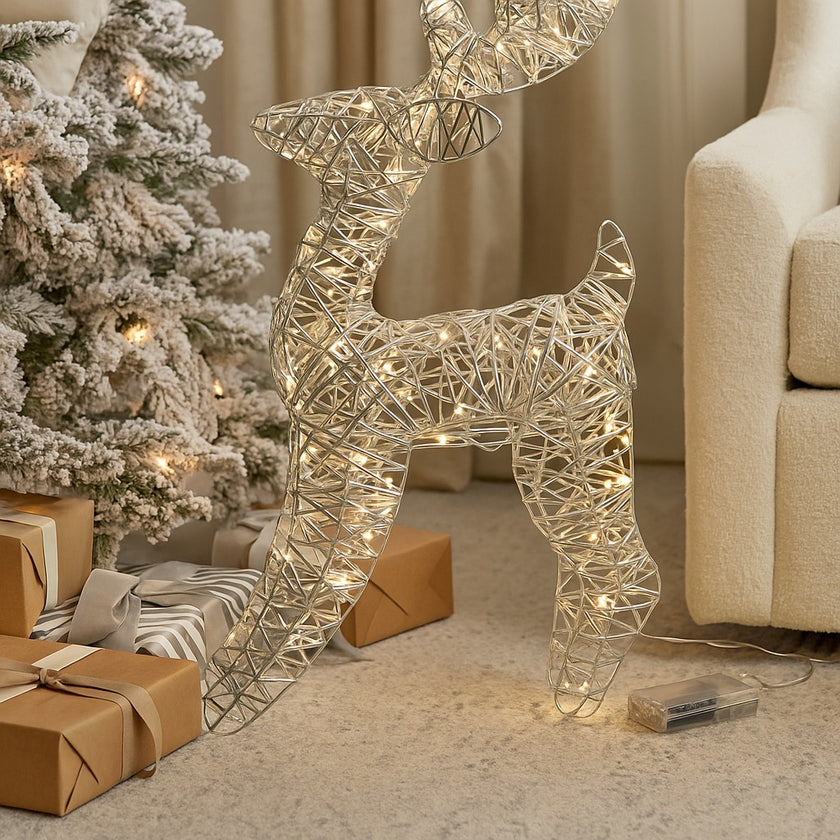 Figurină 3D în formă de ren cu lumini LED așezată în living, lângă un brad nins și cadouri împachetate de Crăciun