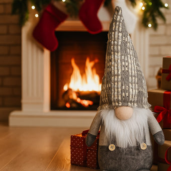 Gnom decorativ de Crăciun cu pălărie lungă gri așezat pe parchet, în fața unui șemineu aprins. Lângă gnom sunt așezate cadouri de Crăciun