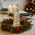 Lumânare în formă de brad auriu cu stele și sclipici așezată în mijlocul unei coronițe de brad, pe o masă de Crăciun