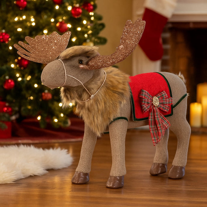 Ren decorativ de Crăciun, din material textil, cu coarne aurii și sclipitoare, așezat pe parchet, în fața unui șemineu și a unui brad decorat de Crăciun