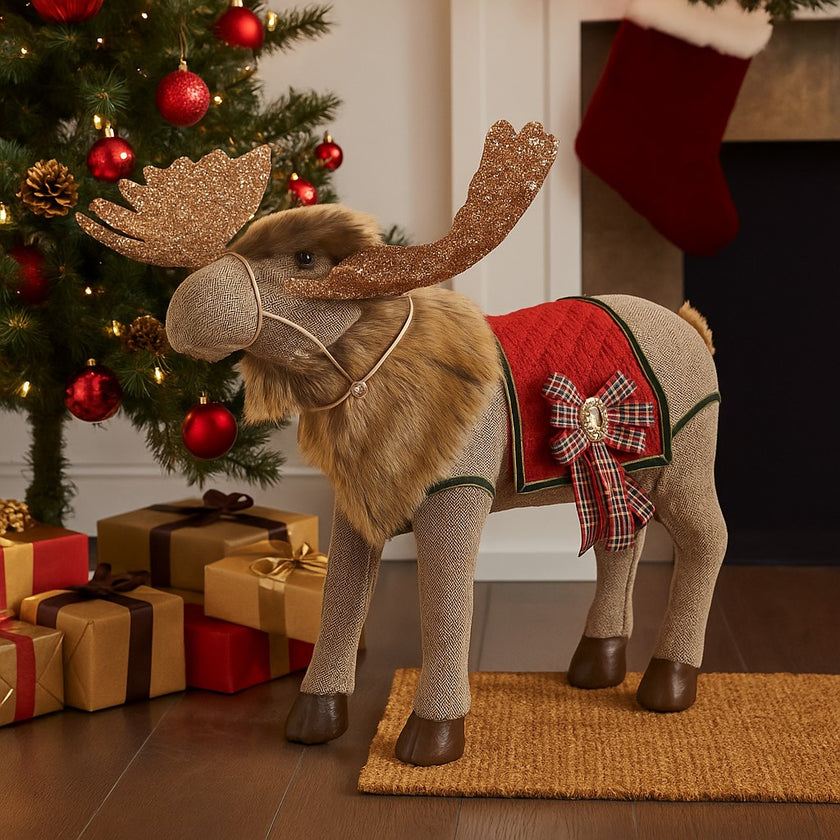 Ren decorativ de Crăciun, din material textil, cu coarne aurii și sclipitoare, așezat lângă un brad decorat de Crăciun sub care se află cadouri împachetate