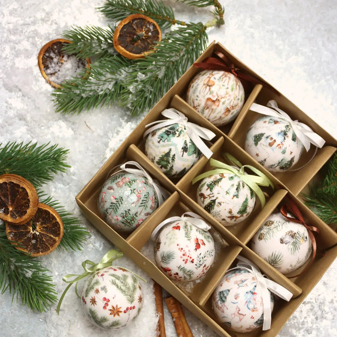 Set globuri de Crăciun cu imprimeuri de iarnă așezate într-o cutie de carton maro. Cutia este așezată pe zapadă