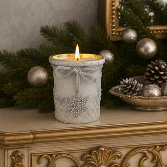 Suport de lumânare din sticlă albă, aprins, pe un bufet decorat de Crăciun cu ramuri de brad, globuri și conuri.