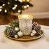 Suport de lumânare din sticlă albă, aprins, pe o tavă aurie decorată de Crăciun cu ramuri de brad, globuri și conuri. Tava este așezată pe un taburete